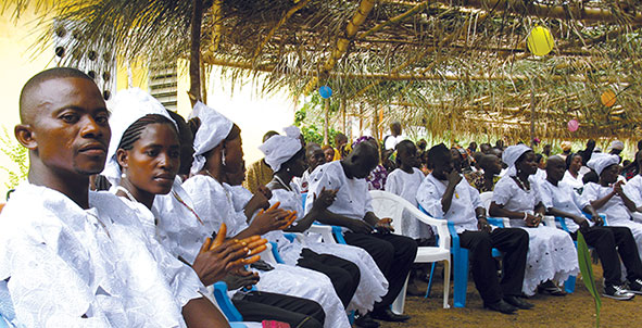 Mariti e mogli seduti su sedie blu e bianche, sotto la tettoia in frasche allestita per l'occasione all'esterno della chiesa San Giuseppe di Marampa, in Sierra Leone.